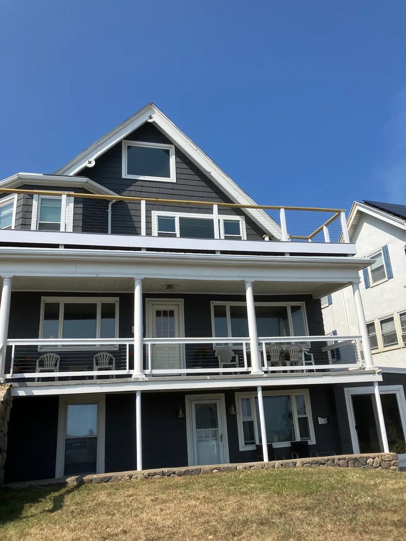 A dark-shingled multi-family house with white trim, an upper deck with cable railings and a covered porch, under a clear blue sky