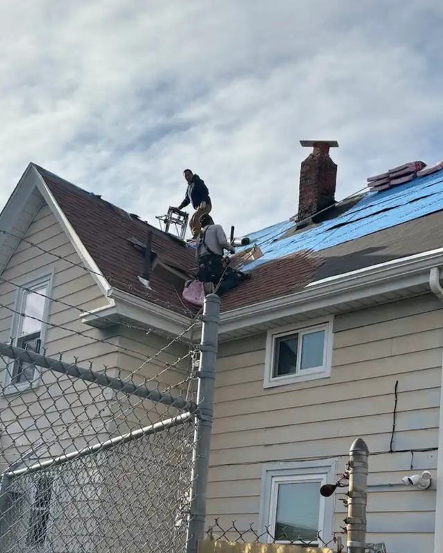 Two roofers stripping old shingles from a two-family house, with blue underlayment going down behind them