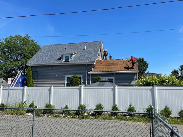 Roofers stripping old shingles from one section of a gray two-story house on a sunny day