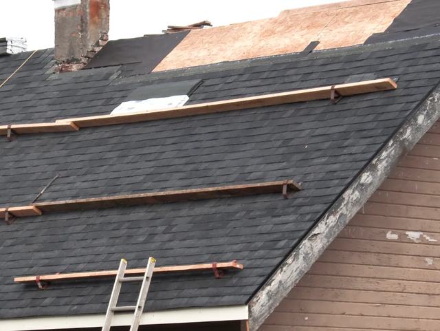 A shingle roof partly opened up to bare sheathing, with roof jacks and a ladder in place