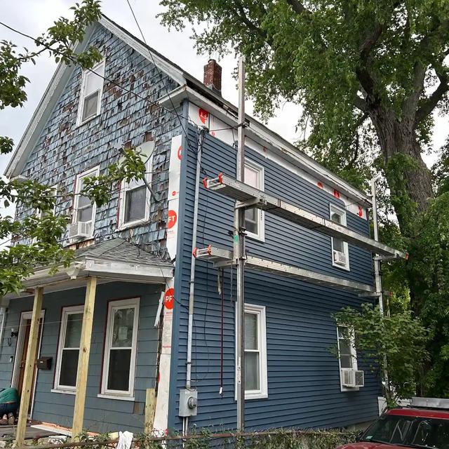 A tall two-family house partway through re-siding, with new blue siding below and old shingles still at the top
