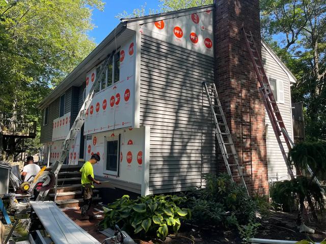 A two-story house wrapped in house wrap with ladders and staging, partway through new gray siding